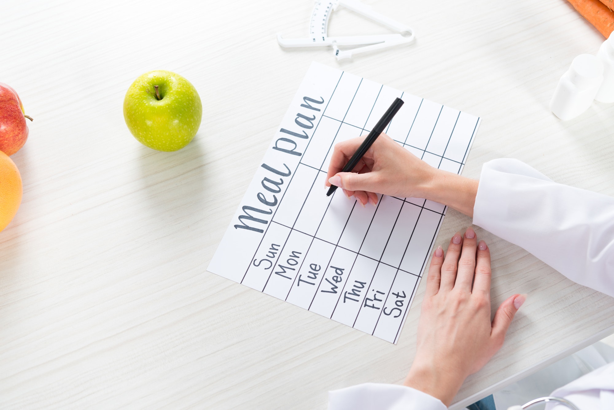 cropped view of dietitian writing in meal plan at table with fruits